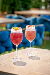 Two vibrant cocktails with foam and garnishes on a wooden table in an outdoor setting with blurred greenery and blue furniture in the background.
