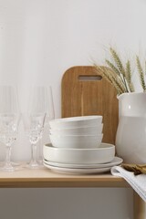 Different clean white dishware, cutlery, cutting board, glasses and spikes on wooden table indoors