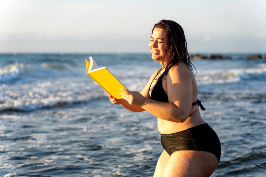 Smiling curvy woman reading a book in the sea waves at sunset
