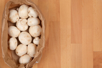 Fresh white mushrooms, or champignons, are shown in a brown paper bag on a wooden table, photographed from above with ample copy space