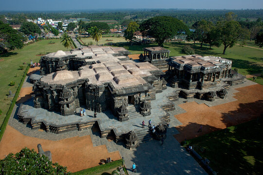 Aerial view of the ancient Kedareshwara Temple's intricate stone carvings and symmetrical architecture contrasting with the surrounding green landscape, Halebeedu, Karnataka, India.
