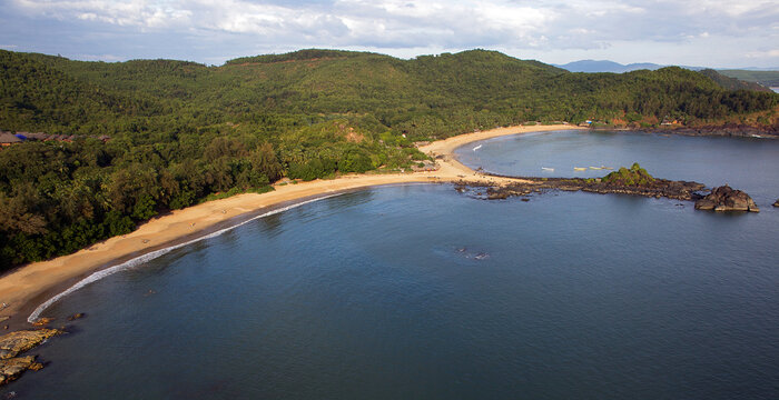 Aerial view of the shoreline where the deep blue sea meets the sandy beach, fringed by verdant forest against a backdrop of rolling hills, Gokarna, Karnataka, India.