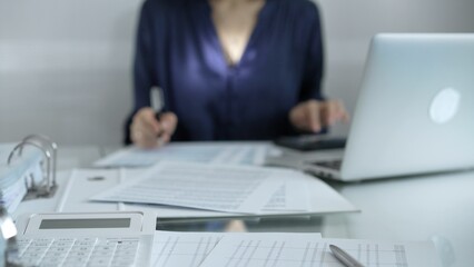 Magnifying glass, white calculator, pen and documents are lying on accountant's desk. Businesswoman wearing blue dress is working with documents on the background. Business and audit concept