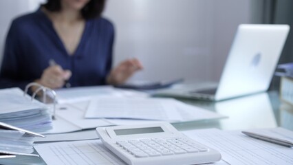 Magnifying glass, white calculator, pen and documents are lying on accountant's desk. Businesswoman wearing blue dress is working with documents on the background. Business and audit concept