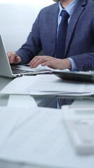 Businessman wearing blue suit is working with laptop, calculator and documents at the glass table in office set. Low lighting, close-up vertical view. Business people and audit