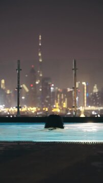 Woman enjoy swimming in a rooftop pool with the stunning and illuminated Dubai city downtown skyline with skyscraper buildings as a backdrop at night