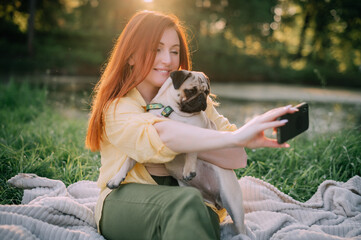 Young redhead woman taking a selfie with her pug dog. Owner and pet having fun outdoors.