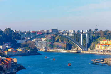 Elegant Ponte da Arrabida spans the Douro River in Porto, framed by colorful riverside buildings and modern apartments. Small boats glide beneath sweeping arch on sunny da