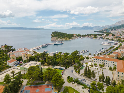 Aerial view of boats resting in a harbor near buildings and lush greenery, with distant mountains meeting the horizon, Makarska, Splitsko-Dalmatinska, Croatia. - Powered by Adobe