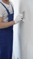 Close-up of man construction worker wearing gloves and blue overalls is using a putty knife to...