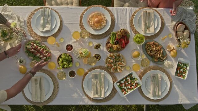 Top view shot of hands putting dishes and decorations on table setup with fresh white tablecloth outdoors in golden hour sunlight, rustic Summer party vibes