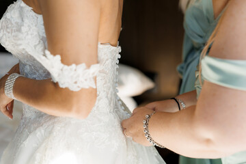 Bridesmaid preparing bride for wedding in room. Bridesmaid helping bride button up wedding dress on...