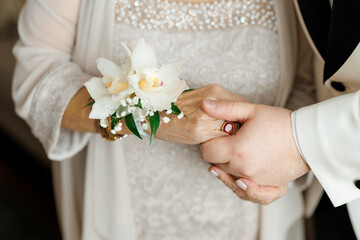Groom holding mother's hand in room on wedding day. Close up of mother's hand with white orchid...