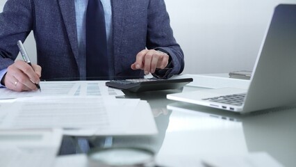 Businessman or accountant wearing blue suit is working with laptop, calculator and documents in office. Low lighting, close-up view. Business people, audit concept