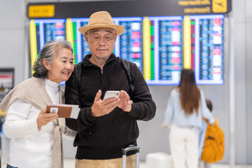 Senior asian couple with luggage checking flight info on smartphone at airport, enjoying retirement...