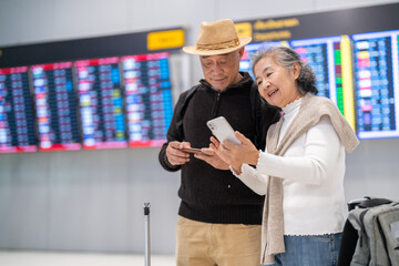 Senior asian couple with luggage checking flight info on smartphone at airport, enjoying retirement...