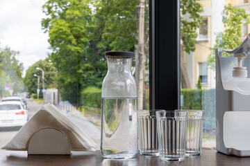 Glass carafe with water, three empty glasses, napkin holder and dispenser on table by window.