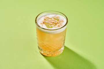 A yellow cocktail with a foam top is garnished with a translucent fruit piece and almond flakes, served in a highball glass on a green background.