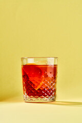 A negroni cocktail with a large, square ice cube and a citrus peel, served in a decorated glass against a yellow background.