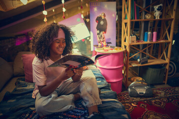 Smiling teen girl sits on bed reading retro magazine in colorful attic with nostalgic decor. Concept of pop culture and music, education and childhood, personal creative space.