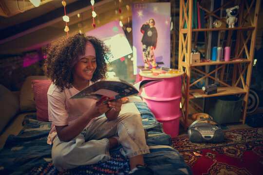 Smiling teen girl sits on bed reading retro magazine in colorful attic with nostalgic decor. Concept of pop culture and music, education and childhood, personal creative space.