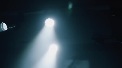 Sharp white spotlights cut through drifting haze in a dark venue, their cones crossing as dust sparkles in the air. The camera lingers on the beams, building a moody, cinematic atmosphere.