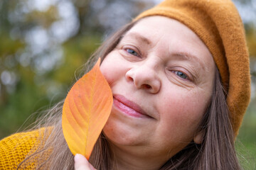 Happy mature woman in beret enjoying autumn nature, with fall leaf in hand, graceful aging, vibrant...