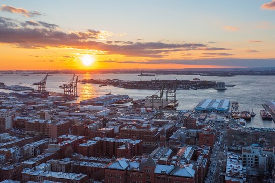 Aerial view of a vibrant sunset painting the sky with hues of orange and gold over the industrial waterfront and cityscape, with the statue of liberty in the background, New York, United States.