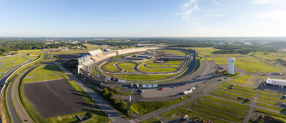 Concord, United States - 28 July 2025: Aerial view of Charlotte Motor Speedway's oval track embraced by lush green fields and winding roads under a vast sky.