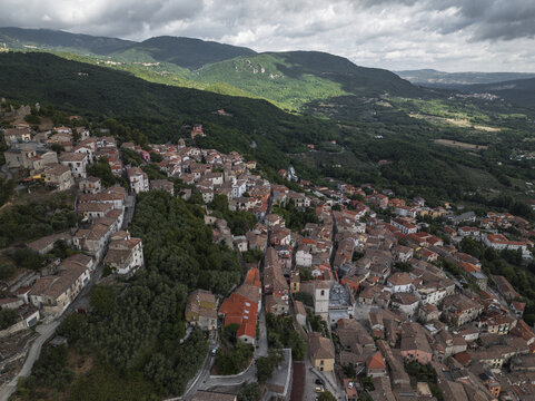Aerial view of clustered terracotta rooftops cascading down the hillside, embraced by verdant slopes under a vast, clouded sky, Morcone, Benevento, Campania, Italy.