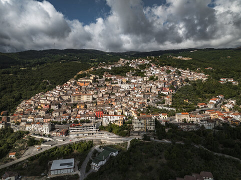 Aerial view of sun-kissed rooftops cascade down the hillside, a tapestry of earthy tones amidst the emerald embrace of the surrounding forests, Morcone, Benevento, Italy.