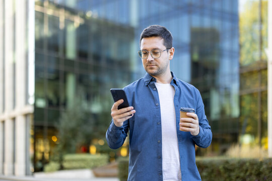 Man with glasses using a smartphone and holding takeaway coffee in an urban setting, blending casual and business attire. The background features modern office buildings, enhancing the corporate vibe - Powered by Adobe