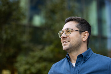 A cheerful man with glasses smiles naturally outdoors, wearing a blue shirt, with blurred green...