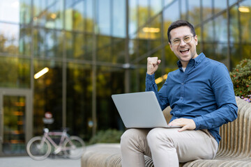 Smiling man sitting outside with a laptop celebrating success, conveying joy and positivity.