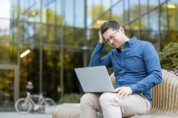 A concerned young man holding his head while working on a laptop outdoors in a modern setting. The...