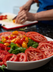 Woman (face not visible) wearing a blue apron and preparing a tomato salad by chopping vegetables including a variety of tomatoes and red onion, and garnishing the dish with basil leaves.