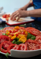 Woman (face not visible) wearing a blue apron and preparing a tomato salad by chopping vegetables including a variety of tomatoes and red onion, and garnishing the dish with basil leaves.
