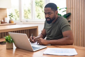 Millennial African American man taking notes during webinar or business meeting, using laptop, working or studying online, sitting at desk in home office. Freelance work, e-commerce startup company.