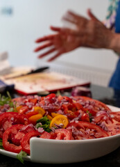 Woman (face not visible) wearing a blue apron and preparing a tomato salad by chopping vegetables including a variety of tomatoes and red onion, and garnishing the dish with basil leaves.