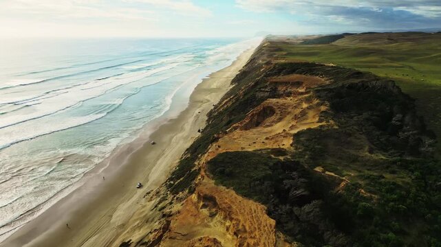 Aerial view of a striking coastline where the turquoise ocean meets a sandy beach, bordered by lush green hills, Dargaville, Northland Region, New Zealand.