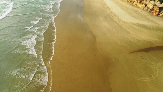 Aerial view of golden sands meeting turquoise waves, contrasted by rugged cliffs topped with green foliage, creating a serene coastal scene, Dargaville, Northland Region, New Zealand.