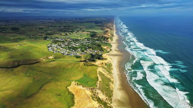 Aerial view of the coastline with a town nestled beside green fields and the ocean's turquoise waves crashing on the sandy shore, Dargaville, Northland Region, New Zealand.