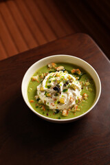 Bowl of green soup on a dark wooden table. The soup is topped with a generous dollop of white cream, surrounded by croutons, chopped green herbs, and small red spices