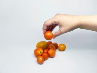A hand delicately holds a single cherry tomato, with a pile of other tomatoes on a clean white surface. The image is well-lit, showcasing the vibrant color and freshness of the produce.