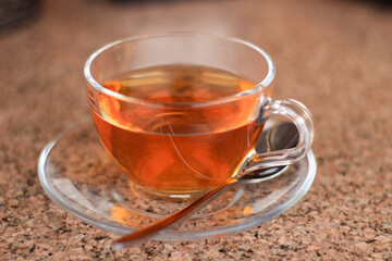 A transparent glass cup filled with amber herbal tea sits on a round glass saucer. A silver spoon rests beside the cup on a textured, light-colored surface.
