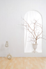 Minimalist white interior with arched window, dried branches in a classic vase, and modern ceramic decor on light wood flooring for a serene and stylish home vibe. design background.