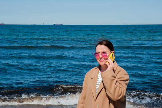 Woman in beige coat and pink sunglasses talking on yellow smartphone while standing on beach near ocean waves under sunny blue sky representing travel and digital nomad lifestyle