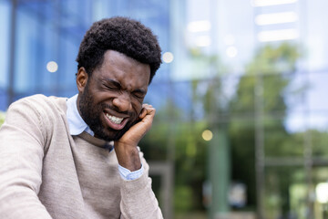 A young man sits outside a glass building, visibly wincing in pain, clutching his jaw clearly...