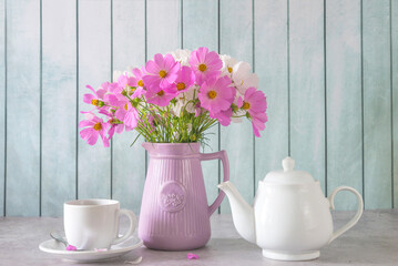 Summer morning still life; Bouquet of pink and white cosmos flowers, white cup of tea or coffee and teapot on a gray table and wooden background