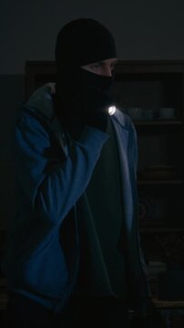 Vertical shot of two criminals with black mask examining living room with flashlight during nighttime